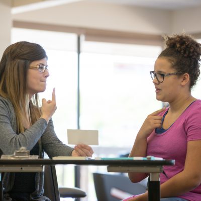 A young adult female speech therapist is helping a teenage patient inside of a clinic